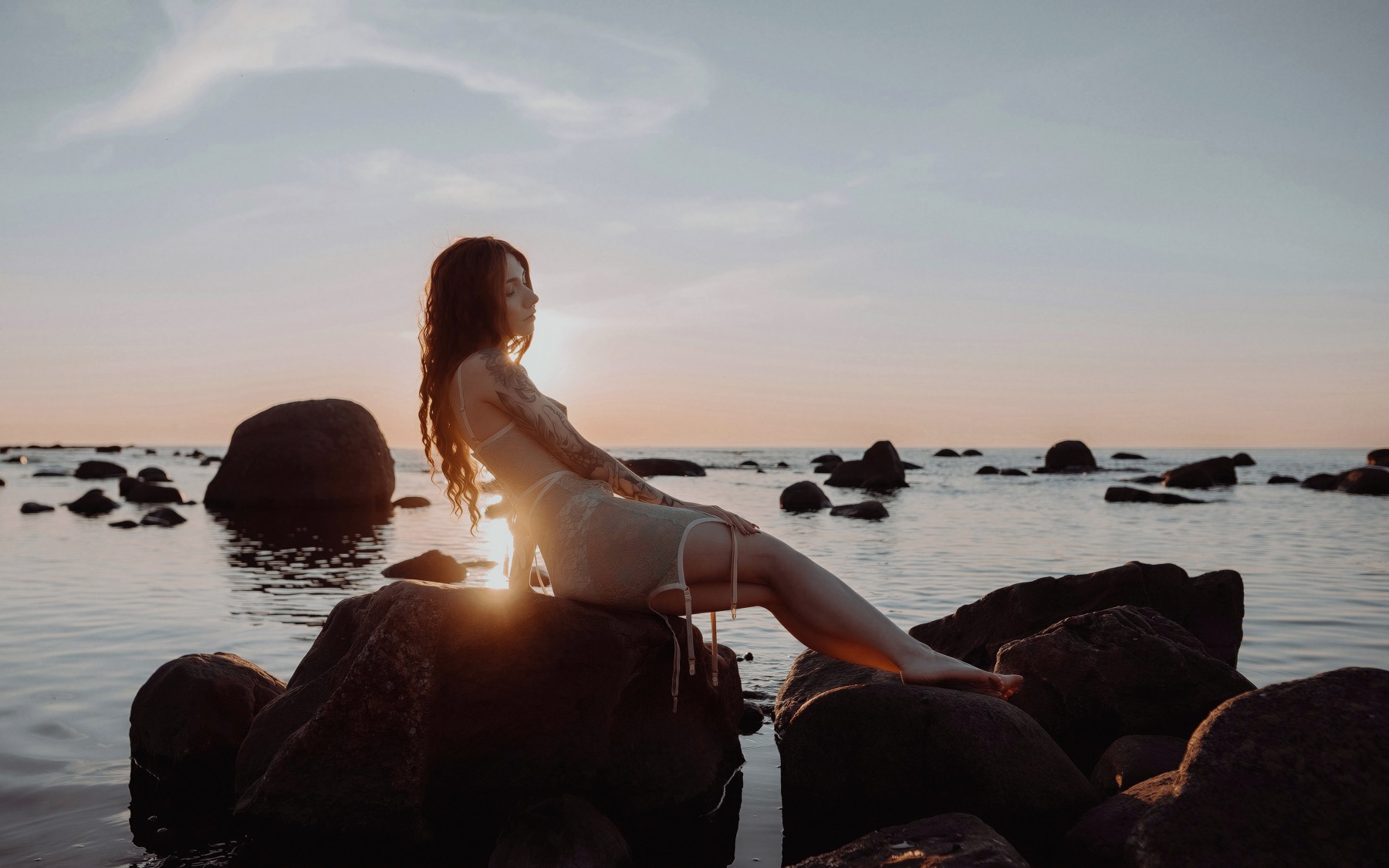 Mulher de cabelos ruivos sobre rochas junto ao mar ao pôr do sol